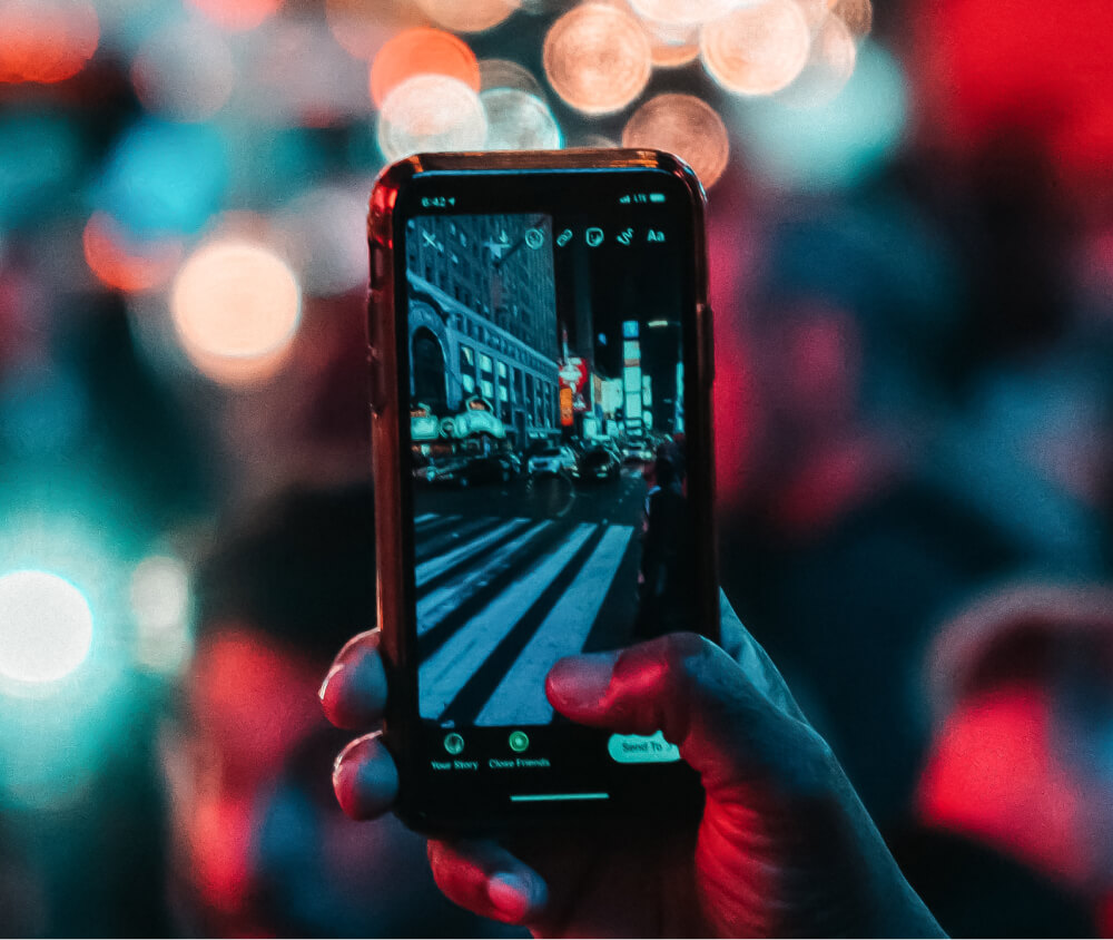 Zbliżenie na dłoń z telefonem. Zdjęcie miasta nocą, w tle rozmazane refleksy światła ulicznych latarni.